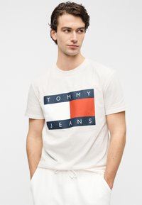 Μπεζ βαμβακερό t-shirt με ορθογώνιο γραφικό σχέδιο που περιλαμβάνει το κείμενο «TOMMY JEANS», σε σκούρο μπλε και κόκκινες λεπτομέρειες. Στυλ με στρογγυλή λαιμόκοψη.