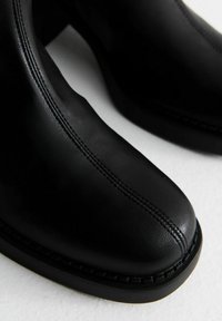 Botas de tobillo de cuero negro con punta redondeada, costuras visibles, diseño elegante y suela de plataforma baja. Textura suave con acentos mínimos.