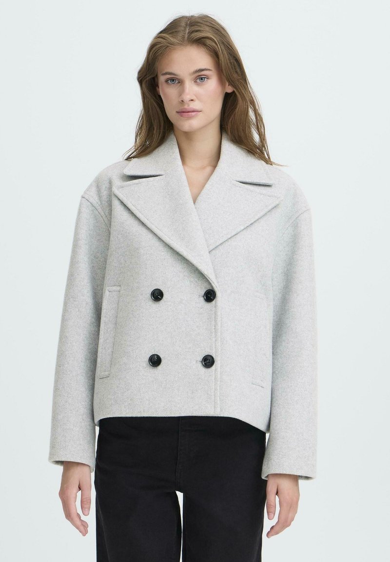 Jeune femme aux cheveux longs, portant un manteau en laine gris clair croisé à double boutonnage et un pantalon noir, se tenant devant un fond clair uniforme.