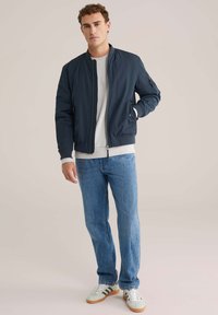 Giubbotto bomber navy con zip frontale, polsini elasticizzati e due tasche laterali, abbinato a jeans azzurri chiari e sneakers grigie con strisce.