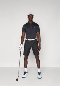 Melns polo krekls ar īsu rāvējslēdzēju, melnas šorti, balts josta, balta golfa cepure un golfa apavi zilu akcentu krāsā. Tur rokā golfa nūju.