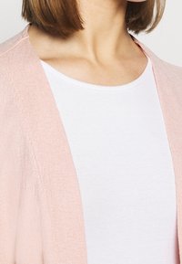 Cárdigan de color rosa claro con una tela texturizada, de frente abierto, combinado con una camisa blanca ajustada. Enfoque en los detalles finos y las costuras.