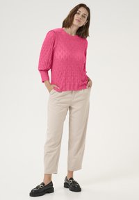 Pull en tricot rose vif avec un motif en diamant, col rond et manches bouffantes, associé à un pantalon beige clair et des mocassins noirs.