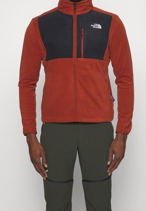 Mann trägt eine rostfarbene Fleecejacke mit schwarzer Brustpartie und Reißverschlusstaschen, kombiniert mit dunkelgrünen Hosen.