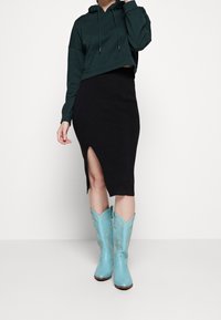 Sudadera corta verde, falda de punto negro con canalé y abertura lateral, y botas vaqueras azules con patrones de costura dorada.