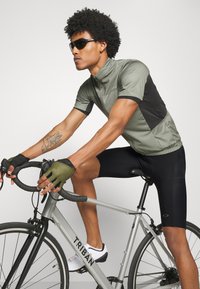 La tenue de cyclisme se compose d'un maillot à manches courtes vert olive clair avec des accents noirs, de shorts noirs et de gants sans doigts. Le vélo est argenté.