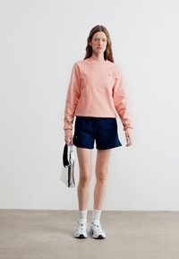 Γυναίκα που στέκεται, φοράει ροζ μακρυμάνικη μπλούζα, σκούρο μπλε σορτς, λευκά αθλητικά παπούτσια και κρατά μια ασημί μεταλλική τσάντα.