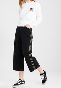 Vit långärmad skjorta med tryck på vänster bröst, svarta vida jeans med guldrand på sidan och svarta Vans sneakers.