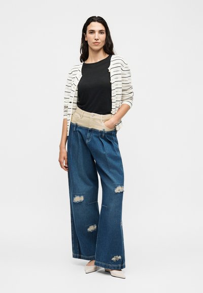Femme debout portant un top noir, un cardigan blanc à motifs, un jean bleu délavé à taille haute et jambes larges, et des chaussures blanches pointues.