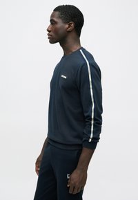 Mladý muž v námornícky modrom tričku s dlhým rukávom a zhodných nohaviciach s bielymi pruhmi s logom na rukávoch a nohaviciach stojí pred jednoduchým svetlým pozadím.