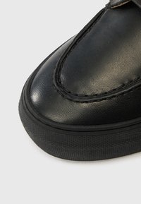 Zapato de cuero negro con acabado liso, punta redondeada y costuras en contraste. Cuenta con una suela de goma negra texturizada para mayor agarre.