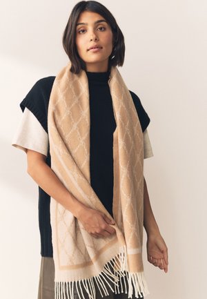 Moteris trumpais tamsiais plaukais, vilkinti juodus ir smėlio spalvos sluoksniuotus drabužius, laikanti smėlio spalvos skarelę su baltomis kutomis.