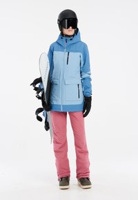 Lichtblauwe waterdichte sneeuwjas met zwarte ritsen, roze snowpants en zwarte handschoenen. Een snowboard wordt onder de arm vastgehouden.