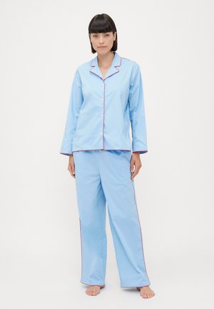 Lyseblåt pyjamassæt bestående af en skjorte med knapper og lange ærmer samt matchende bukser, kantet med rød piping langs kraven og sømmene.