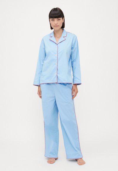 Lyseblåt pyjamassæt bestående af en skjorte med knapper og lange ærmer samt matchende bukser, kantet med rød piping langs kraven og sømmene.