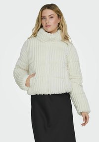 Giubbotto imbottito bianco con collo alto, caratterizzato da una texture a costine, chiusura con zip e tasche laterali. Indossato sopra una gonna scura.