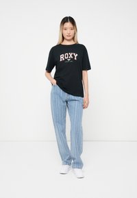 Must mustaga, millel on valge ja punane "ROXY" logo, koos heledate siniste triibuliste teksade ja valgete kingadega; seisev poos tasasel taustal.