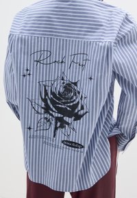Camicia a righe blu e bianche con una grande grafica di una rosa nera e testo sulla schiena, con orlo arrotondato e tessuto morbido.
