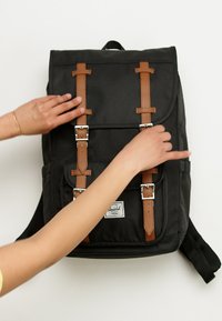 Crni platneni ruksak s smeđim kožnim remenjem i srebrnim metalnim detaljima. Ima zatvaranje na preklop i podesive naramenice.