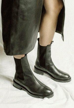 Black leather ankle boots with elastic side panels, rounded toes, and textured rubber soles, paired with a black leather skirt.