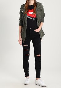 Kvinna i grön hoodiejacka, svart t-shirt med grafiskt tryck av röda läppar, sönderrivna svarta slim-fit-jeans och vita sneakers, stående med en hand i fickan.