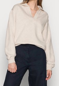 Pull-over beige à manches longues avec col en V et poignets côtelés, associé à un pantalon foncé. Tissu doux et texturé, coupe décontractée.