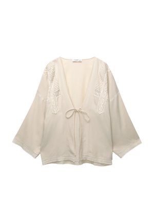Blusa stile kimono color beige chiaro con maniche ampie, scollatura a V profonda e raffinato pizzo decorativo lungo le spalle e l'orlo. Legata in vita.