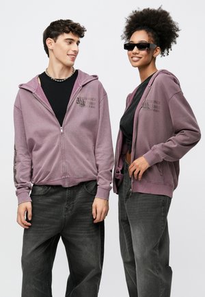 Lilla hettejakke med glidelås og logo på forsiden, kombinert med svarte topper og mørke jeans. Den kvinnelige modellen har på seg solbriller, begge plaggene har en avslappet passform.