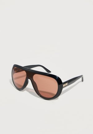 Ochelari de soare supradimensionați negri cu lentile maro și ramă groasă, așezați înclinați pe o suprafață albă.