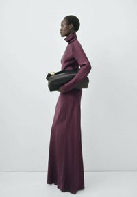 Long, fitted burgundy dress with a high neck, made of soft fabric. Model holds a sleek black bag with a rounded gold clasp.