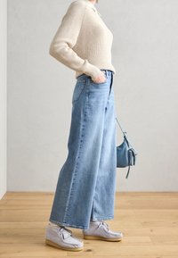 Beige geribde trui, hoogwaist lichtblauwe wijde jeans, grijze schoenen met veters, met een kleine lichtblauwe handtas.