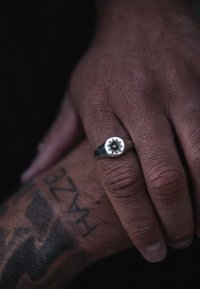 Anello d'argento con un design circolare a raggi di sole che presenta una piccola pietra centrale. La fascia è liscia e lucida, indossata su una mano con texture.
