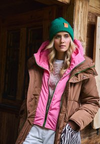 Brązowa kurtka puchowa z różowym wnętrzem, zielonymi zamkami błyskawicznymi i zieloną dzianinową czapką. Widoczny detal kieszeni w wzór zebry.