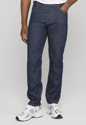Homme portant un jean bleu foncé et des baskets blanches New Balance, debout devant un fond clair uni.
