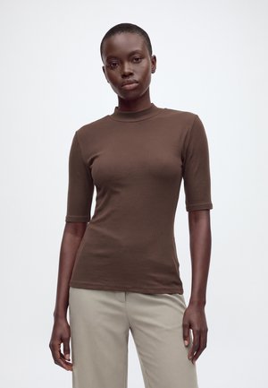 Un modelo posa sobre un fondo blanco liso, vistiendo una camiseta ajustada de manga corta marrón con textura de canalé y pantalones beige.