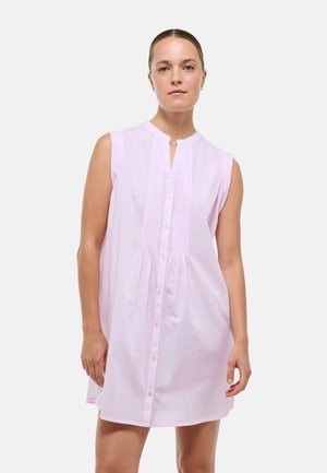 Femme portant une robe sans manches rose clair avec des boutons sur le devant et un détail plissé à l’avant, debout devant un fond clair uni.