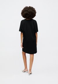 Black oversized t-shirt dress with short sleeves, smooth texture, and a rounded hem. Paired with light gray high-heeled shoes.