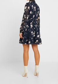 En kvinna i en blommig marinblå klänning med långa ärmar och en volangkant står med ryggen mot kameran, iförd krämfärgade ankle boots.