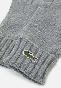 Grå ribbestrikkede hansker laget av mykt materiale, med en liten brodert grønn krokodille-logo med røde detaljer på mansjetten.