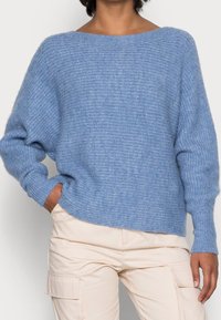 Pull bleu clair tricoté avec un large col, épaules tombantes et poignets côtelés. Présente une finition douce et texturée ainsi qu'une coupe décontractée.