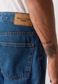 Nærbilde av baklommen på blå denimjeans og lærmerket til Marc O'Polo Denim, med en tatovert underarm som hviler ved siden av.