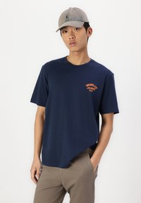 Námornícky modré bavlnené tričko s krátkymi rukávmi a okrúhlym výstrihom, zdobené oranžovým logom "Wrangler Jeans 1947" na hrudi.