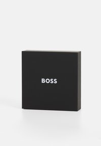 Boîte rectangulaire noire en matériau mat avec une texture lisse. Présente un texte en relief blanc "BOSS" sur le devant.