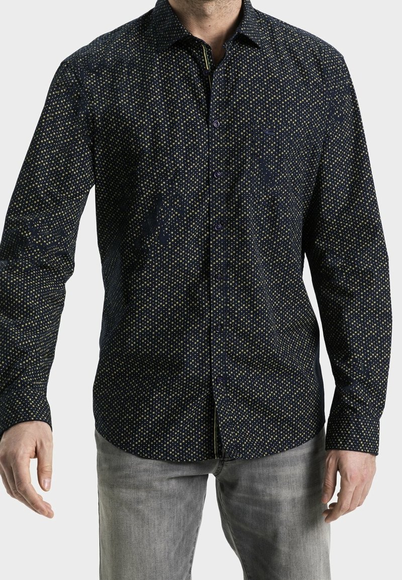 Homme portant une chemise bleu marine ajustée avec un petit motif carré jaune et un jean gris clair, debout devant un fond uni.