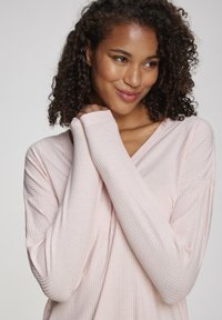 Light pink long-sleeve top with a V-neck, featuring a ribbed texture and extended cuff details. Soft fabric drapes gently.