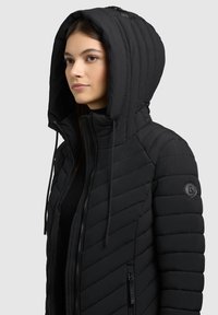 Kobieta nosząca czarną pikowaną kurtkę z kapturem, zapinaną na suwak z przodu i z naszywką z logo na rękawie, zwrócona w lewo na jednolitym tle.
