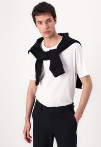White t-shirt paired with dark trousers, featuring a black sweater draped over the shoulders, displaying a casual, layered look.