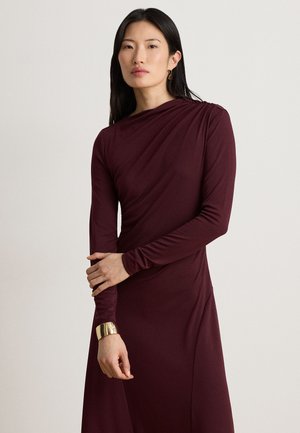 Frau mit langen dunklen Haaren, die ein langärmliges burgunderrotes Kleid und ein goldenes Armband trägt, steht vor einem schlichten hellen Hintergrund.