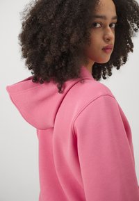 Felpa rosa realizzata in tessuto testurizzato, presenta un ampio cappuccio, polsini a costine e una vestibilità rilassata, indossata da un modello con capelli ricci.