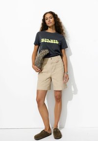 T-shirt noir avec motif jaune, short beige et chaussures slip-on vert olive. Tenant une pochette imprimée léopard. Fond blanc.